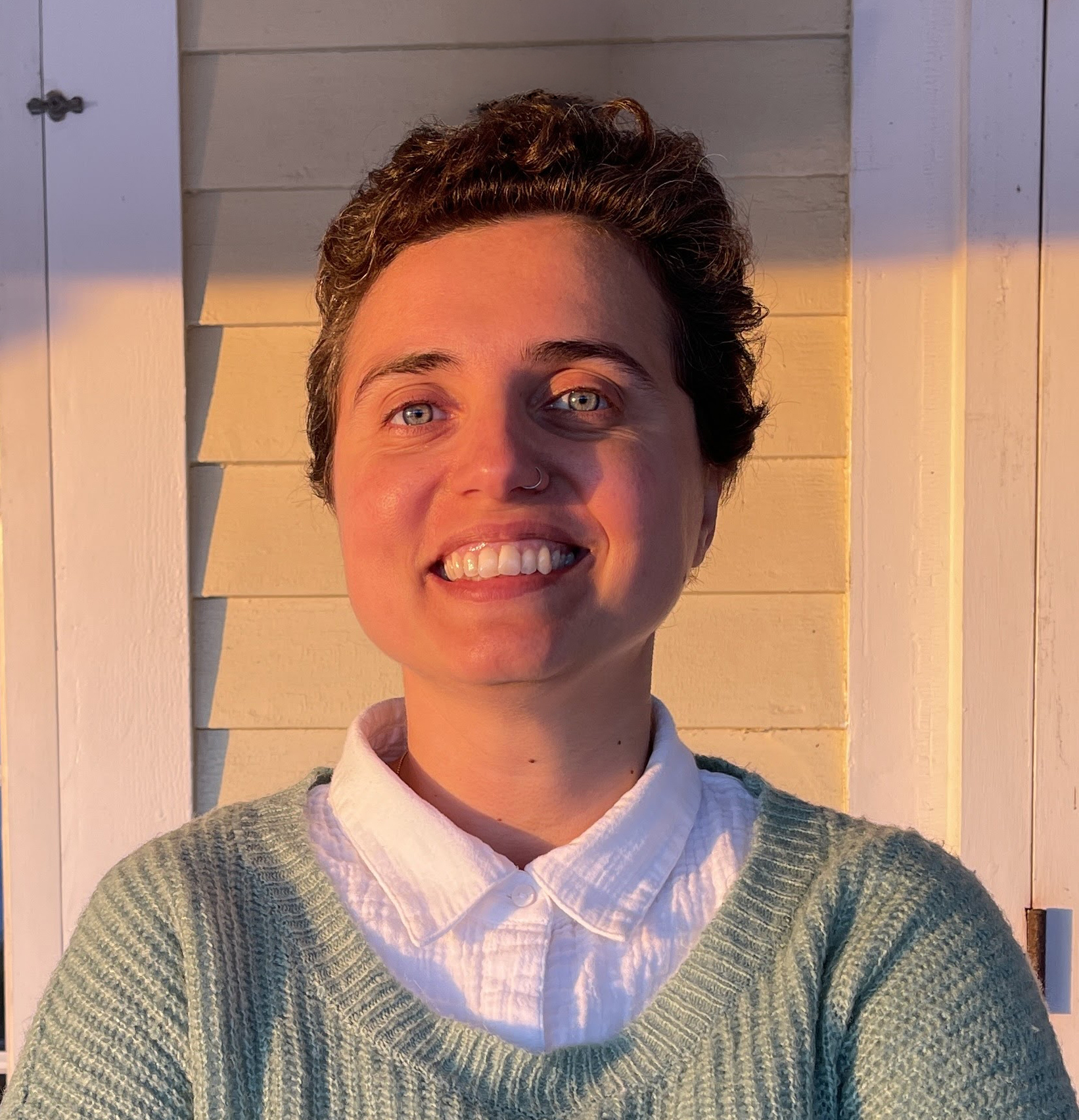 A woman with short hair and white shirt and mint-green sweater smiling into the camera. She is in front of a yellow wall.